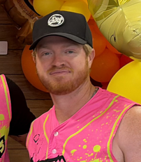 Dustin Baber Man with red hair and beard wearing a black cap and a bright pink shirt.