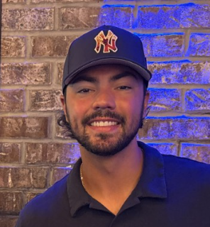 Jace Avina Young man with a beard wearing a baseball cap, smiling in front of a brick wall.